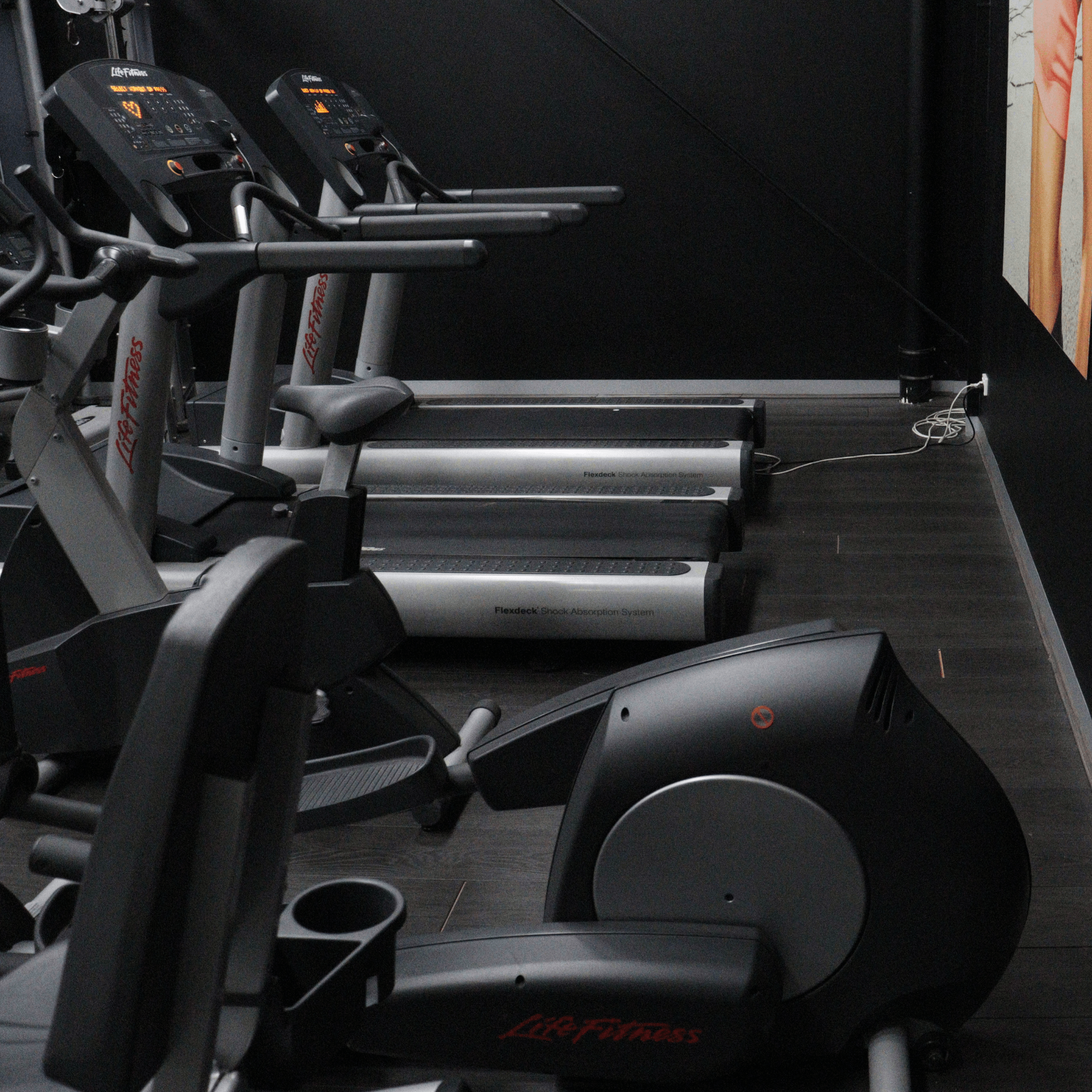 Gym equipment including treadmills and exercise bikes with a dark floor and wall.